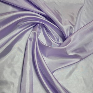 May include: A close-up shot of a light purple fabric with a satin sheen. The fabric is draped and folded, creating soft shadows and highlights. The material appears smooth and flowing, suggesting a lightweight textile.