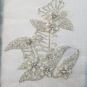 May include: A silver and white beaded floral applique with a large central flower and smaller flowers on the sides. The applique is approximately 5 inches wide and 6 inches tall.