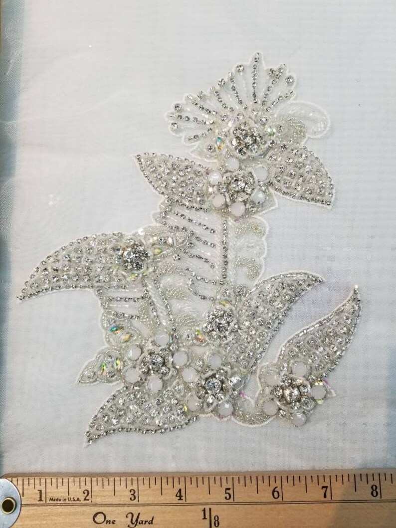 May include: A silver and white beaded floral applique with a large central flower and smaller flowers on the sides. The applique is approximately 5 inches wide and 6 inches tall.