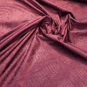 Może przedstawiać: Zbliżenie na burgundowy materiał z tłoczonym wzorem krokodyla. Materiał jest miękki i fakturowany.