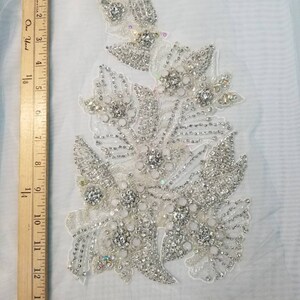 Peut inclure: Applique florale blanche orn&eacute;e de perles et de strass scintillants. Mesure environ 15 cm de large et 25 cm de long. Le motif pr&eacute;sente diverses formes de fleurs et de feuilles, id&eacute;al pour les tenues de mari&eacute;e ou de c&eacute;r&eacute;monie.