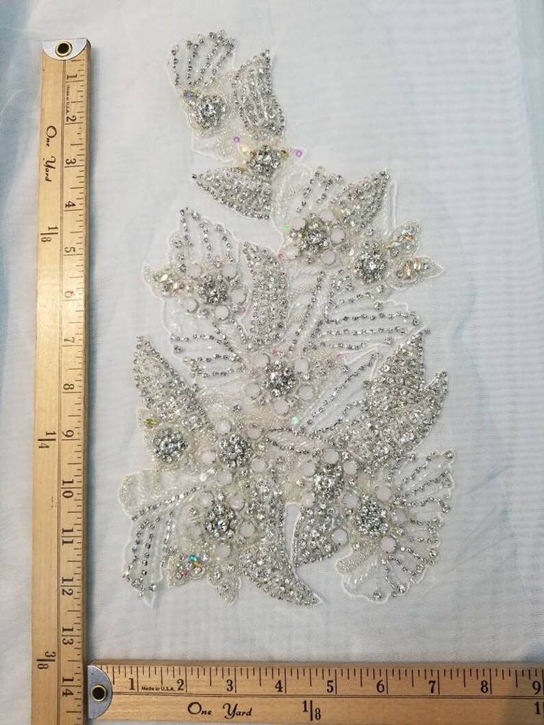 Peut inclure: Applique florale blanche orn&eacute;e de perles et de strass scintillants. Mesure environ 15 cm de large et 25 cm de long. Le motif pr&eacute;sente diverses formes de fleurs et de feuilles, id&eacute;al pour les tenues de mari&eacute;e ou de c&eacute;r&eacute;monie.