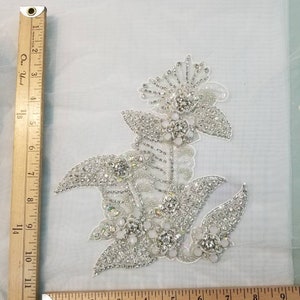 May include: A silver and white floral applique with rhinestones and pearls. The applique is approximately 4 inches wide and 5 inches tall.