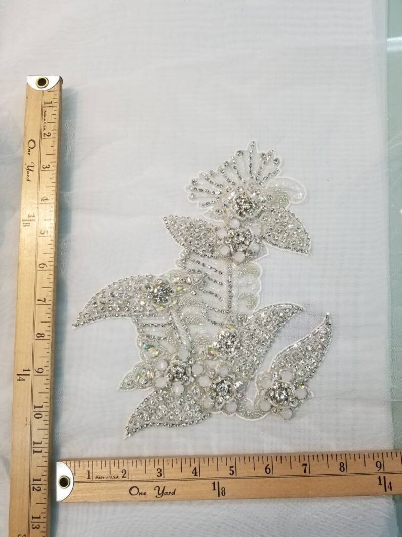 May include: A silver and white floral applique with rhinestones and pearls. The applique is approximately 4 inches wide and 5 inches tall.