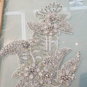 May include: A delicate floral design with white and silver embellishments. The design features a large flower with smaller flowers and leaves surrounding it. The embellishments are made of rhinestones and beads.