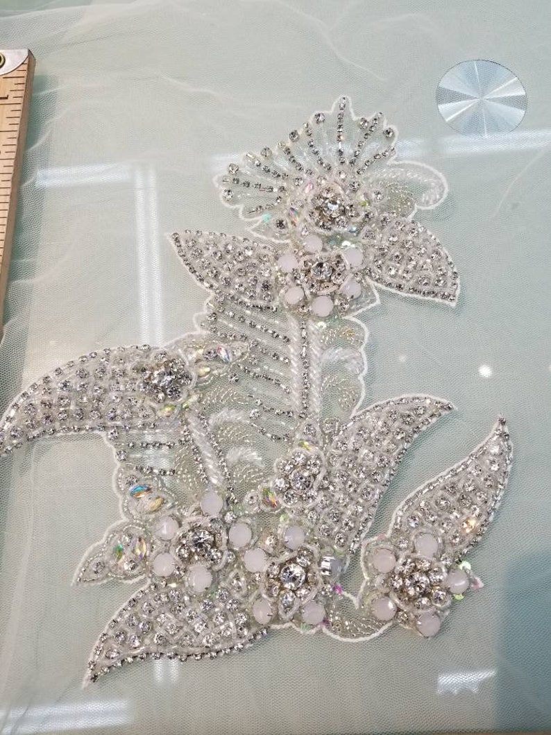 May include: A delicate floral design with white and silver embellishments. The design features a large flower with smaller flowers and leaves surrounding it. The embellishments are made of rhinestones and beads.
