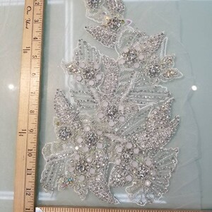 Peut inclure: Une applique florale argent&eacute;e d&eacute;taill&eacute;e avec des strass transparents et iris&eacute;s et des perles blanches. L'applique est sur un tissu transparent de couleur claire. Une r&egrave;gle en bois est dans l'image.