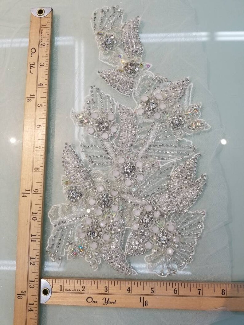 Peut inclure: Une applique florale argent&eacute;e d&eacute;taill&eacute;e avec des strass transparents et iris&eacute;s et des perles blanches. L'applique est sur un tissu transparent de couleur claire. Une r&egrave;gle en bois est dans l'image.