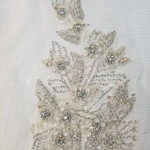Peut inclure: Applique florale d&eacute;taill&eacute;e, avec une base blanche orn&eacute;e de strass et de perles. Le motif pr&eacute;sente des groupes de cristaux clairs entour&eacute;s de petites perles argent&eacute;es, cr&eacute;ant un effet scintillant. Id&eacute;al pour embellir les robes de mari&eacute;e ou de soir&eacute;e.