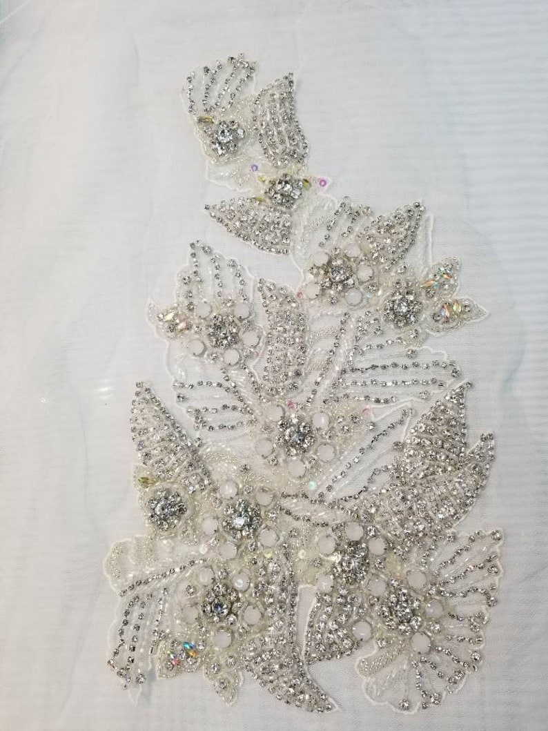 Peut inclure: Applique florale d&eacute;taill&eacute;e, avec une base blanche orn&eacute;e de strass et de perles. Le motif pr&eacute;sente des groupes de cristaux clairs entour&eacute;s de petites perles argent&eacute;es, cr&eacute;ant un effet scintillant. Id&eacute;al pour embellir les robes de mari&eacute;e ou de soir&eacute;e.