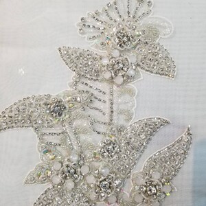 May include: A white fabric applique with a floral design made of silver and white beads and rhinestones.