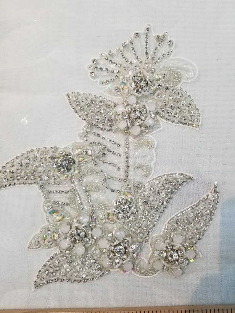 May include: A white fabric applique with a floral design made of silver and white beads and rhinestones.