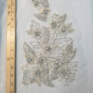 Peut inclure: Applique florale d&eacute;taill&eacute;e orn&eacute;e de strass et de perles transparents et iris&eacute;s. L'ornement est pos&eacute; sur un tissu blanc transparent. L'applique mesure environ 33 cm de long.