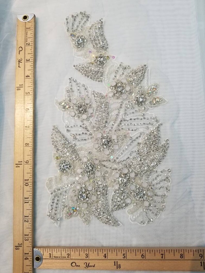 Peut inclure: Applique florale d&eacute;taill&eacute;e orn&eacute;e de strass et de perles transparents et iris&eacute;s. L'ornement est pos&eacute; sur un tissu blanc transparent. L'applique mesure environ 33 cm de long.