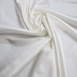 May include: A close-up of a white fabric with a smooth, silky texture. The fabric is draped and folded, creating soft, flowing lines.