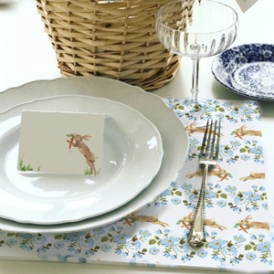 May include: A wicker basket filled with paper, a bottle of champagne, and a chocolate bunny. A clear champagne glass, white plates, and a floral placemat with a fork are on the table. A card with a bunny illustration is on a plate.