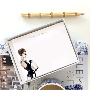 May include: A box of personalized stationery featuring a black and white illustration of a woman in a black dress and sunglasses. The box is white with a silver trim and the name "SAMANTHA SMITH" is printed on the front.
