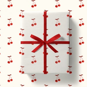 May include: A wrapped gift with white paper featuring a pattern of red cherries. The gift is tied with a red satin ribbon and bow. The background is a solid off-white color.