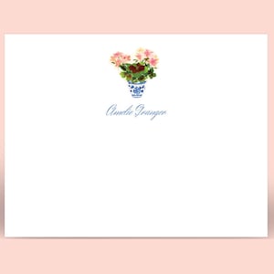 May include: White stationery with a watercolor illustration of pink flowers in a blue and white vase. The name "Amelie Granger" is written in blue script below the vase. The background is a soft pink.