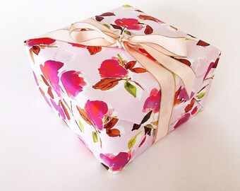 May include: A wrapped gift box with a floral pattern in shades of pink, red, and purple. The box is tied with a light pink ribbon with gold trim, creating a decorative and elegant presentation.