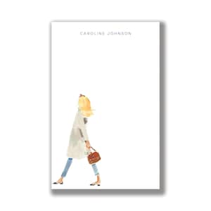 May include: A white notepad with the name "CAROLINE JOHNSON" in blue at the top. A watercolor illustration of a person in a white coat, blue jeans, and carrying a brown purse is on the notepad. The person has blonde hair and is walking.