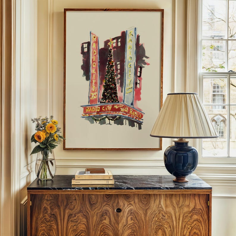 Op de afbeelding: Een aquarel van Radio City Music Hall in New York City met een kerstboom voor het gebouw. Het schilderij is ingelijst en hangt aan een muur boven een houten kast.
