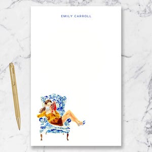 May include: White notepad with the text "EMILY CARROLL" in blue. The notepad features a watercolor illustration of a person reading in a blue and white patterned chair. A gold pen rests on the left.