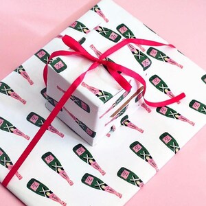 May include: White gift wrap and a small box feature a pattern of green and pink champagne bottles. A bright red ribbon is tied around the box. The background is a solid pink color.