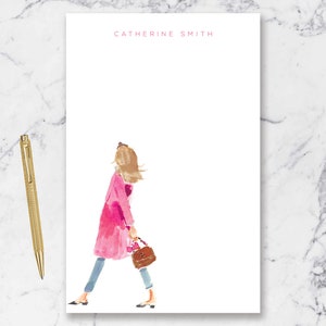 May include: A pink notepad with the name "Catherine Smith" printed at the top. A watercolor illustration of a woman in a pink coat walking with a brown handbag is on the notepad.