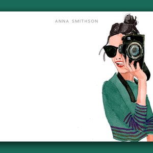 May include: A watercolor illustration of a woman wearing sunglasses and a teal sweater with a striped pattern. She is holding a black camera and smiling.