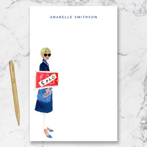 May include: A white notepad with a blue and red illustration of a woman holding a red "SOLD" sign. The notepad has the name "ANABELLE SMITHSON" printed at the top.