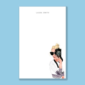 May include: Illustration of a notepad with the name "LAURA SMITH" at the top. A stylish illustration of a person wearing sunglasses and a striped shirt holding a black camera. The background is a light blue color.
