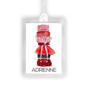 May include: A clear luggage tag with a pink and red illustration of a woman carrying suitcases. The text "ADRIENNE" is printed on the tag.