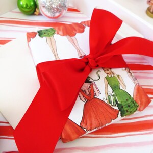 Puede incluir: Una cinta roja atada alrededor de una caja de regalo blanca con un patr&oacute;n de rayas rojas y blancas. La caja de regalo est&aacute; envuelta en papel con un dise&ntilde;o de mujeres con vestidos.