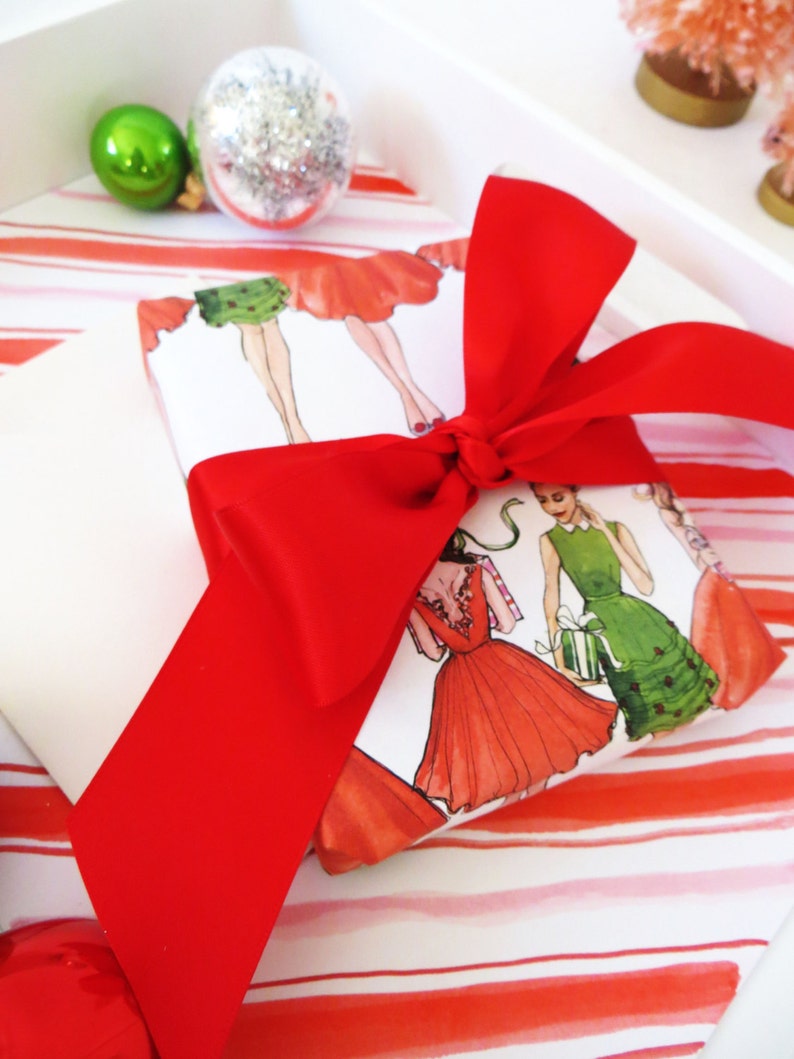 Puede incluir: Una cinta roja atada alrededor de una caja de regalo blanca con un patr&oacute;n de rayas rojas y blancas. La caja de regalo est&aacute; envuelta en papel con un dise&ntilde;o de mujeres con vestidos.