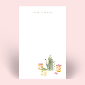 May include: A white notepad with the name "SUSAN THORNTON" at the top. The bottom of the notepad features a watercolor illustration of a silver tea pot and three decorative boxes. The background is a soft pink.