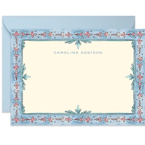 May include: A light blue stationery set featuring a cream-colored card with a decorative border of pink and teal floral elements. The card includes the name "CAROLINE ADDISON" in a matching blue font. A light blue envelope is behind the card.