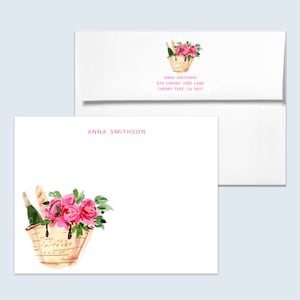 May include: A white card with pink text that reads "Anna Smithson". The card features a watercolor illustration of a wicker basket filled with pink flowers, a bottle of champagne, and bread.