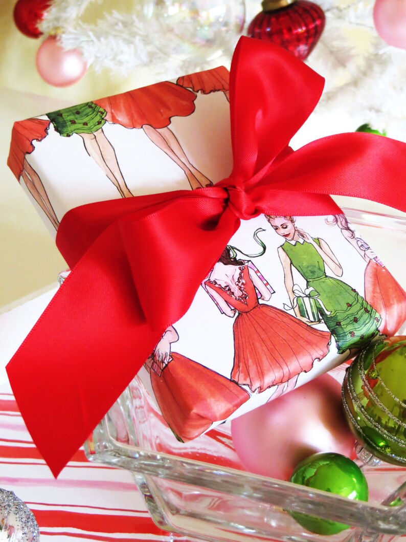 Puede incluir: Una caja de regalo blanca envuelta con una cinta de sat&eacute;n roja y decorada con un patr&oacute;n de mujeres con vestidos. La caja est&aacute; sobre una superficie de vidrio transparente con adornos verdes y rosas.
