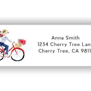 May include: A white label with a watercolor illustration of a person riding a red bicycle with a basket of flowers. The label includes the name "Anna Smith" and an address.