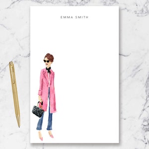 May include: A white notepad with the name "EMMA SMITH" printed at the top. A watercolor illustration of a woman wearing a pink coat, sunglasses, and a black scarf is on the left side of the notepad.