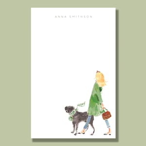 May include: A white notepad with the name "ANNA SMITHSON" printed at the top. The notepad features a watercolor illustration of a woman in a green coat walking a black dog with a green scarf.