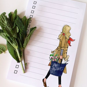 May include: A white notepad with a lined interior and a checklist on the left side. The notepad features a hand-drawn illustration of a woman in a tan trench coat carrying a blue tote bag with the text "Le Market" on it. The woman is wearing a red and white patterned scarf.
