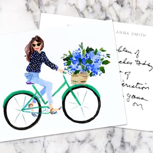 K&ouml;nnte beinhalten: Aquarellillustration einer Frau auf einem gr&uuml;nen Fahrrad mit einem Korb voller blauer Blumen. Die Frau tr&auml;gt eine Sonnenbrille, ein marineblaues Polka-Dot-Hemd und Jeans. Ein goldener Stift und eine Karte mit handschriftlichem Text sind ebenfalls zu sehen.