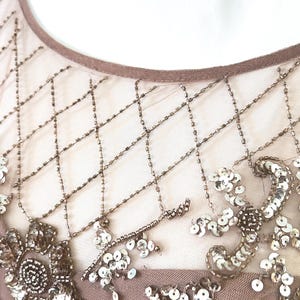 May include: Close-up of a formal dress bodice. The sheer overlay features a diamond pattern created with small, bronze-colored beads. Floral and leaf designs are embellished with sequins and beads in shades of silver and bronze.
