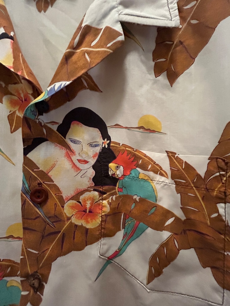 May include: A white button-up shirt with a tropical print featuring a woman, a parrot, and brown palm leaves. The shirt has a pocket on the left side.