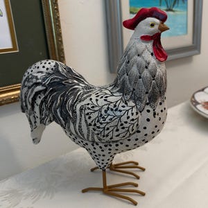 May include: A decorative rooster figurine featuring a white body with black polka dots and intricate black line patterns on the wings and tail. The rooster has a bright red comb and wattle, a yellow beak, and thin, brown metal legs and feet.