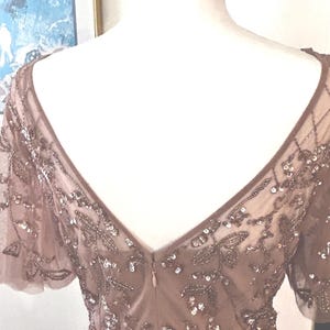 May include: A mauve-colored formal dress with a V-neckline and short sleeves. The dress is adorned with intricate beadwork and sequins, creating a shimmering effect. The back of the dress features a low V-cut and a zipper closure.