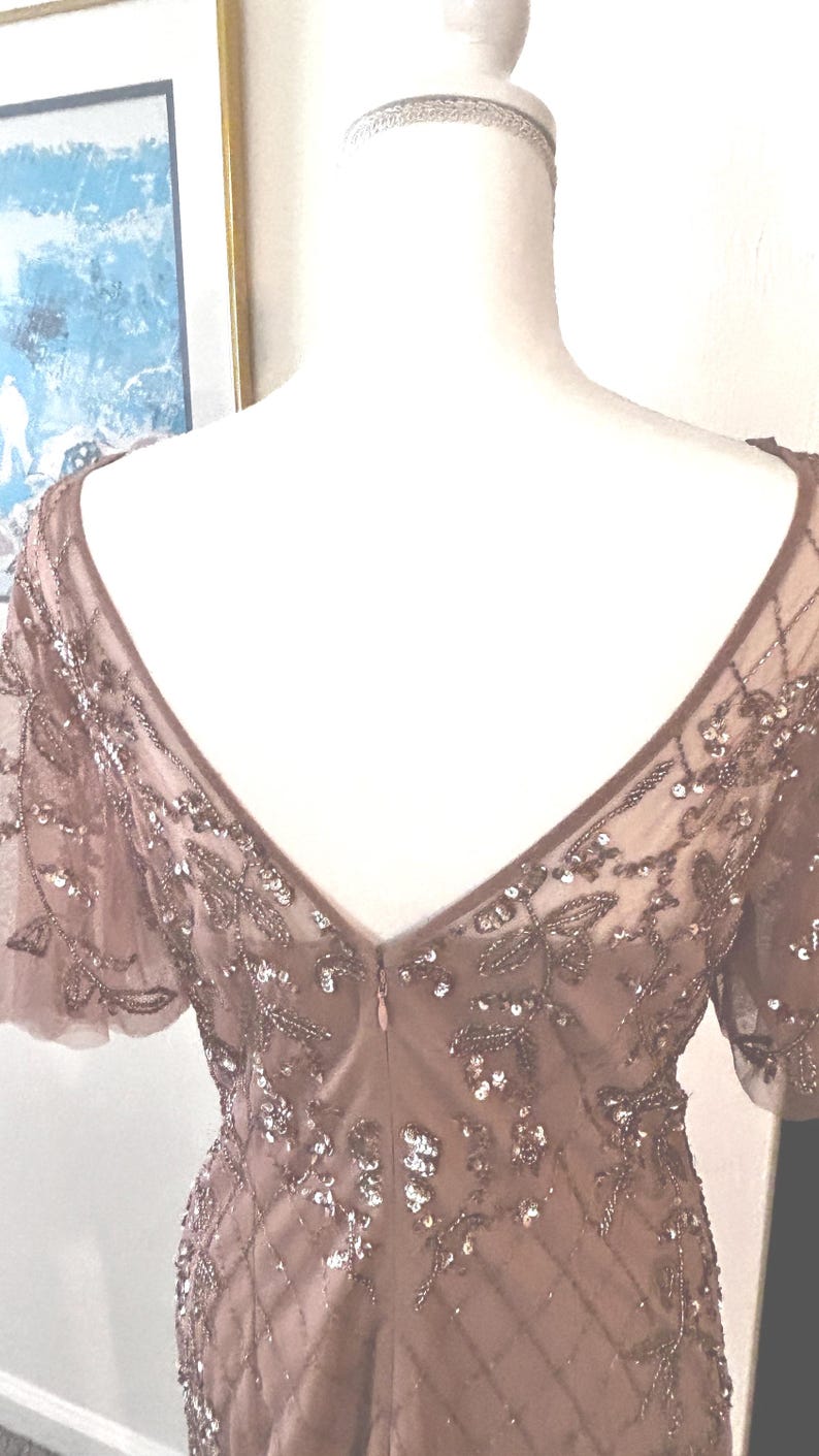 May include: A mauve-colored formal dress with a V-neckline and short sleeves. The dress is adorned with intricate beadwork and sequins, creating a shimmering effect. The back of the dress features a low V-cut and a zipper closure.