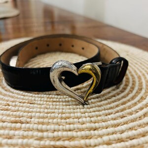 May include: Black belt with a silver and gold heart-shaped buckle. The belt is made of a textured material and has a classic design. The buckle is a prominent feature, adding a touch of elegance to the accessory.